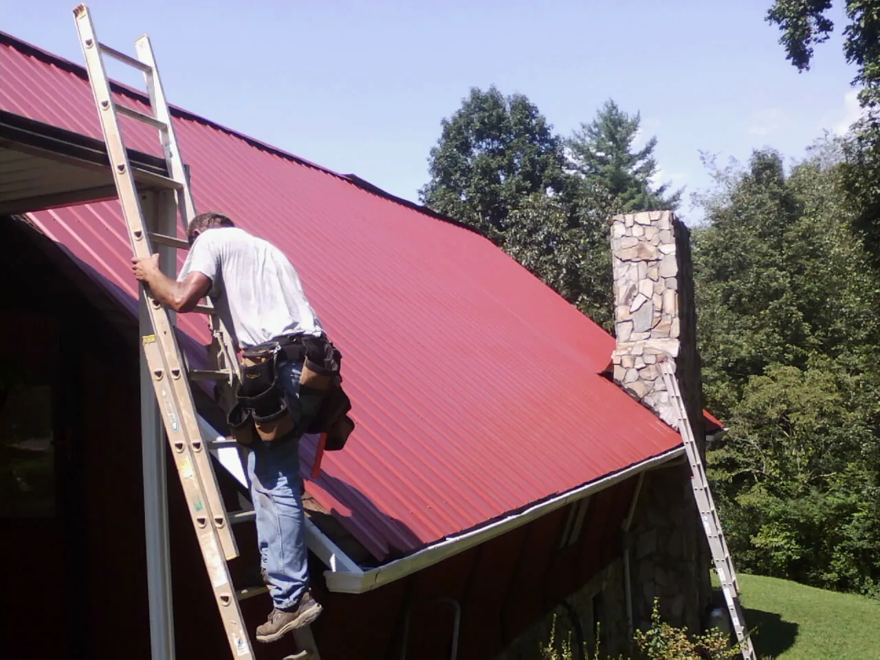 Roofer on a ladder beside a red metal roof and stone chimney — Soffit & Fascia Repair by Roofing Brothers in Shiro